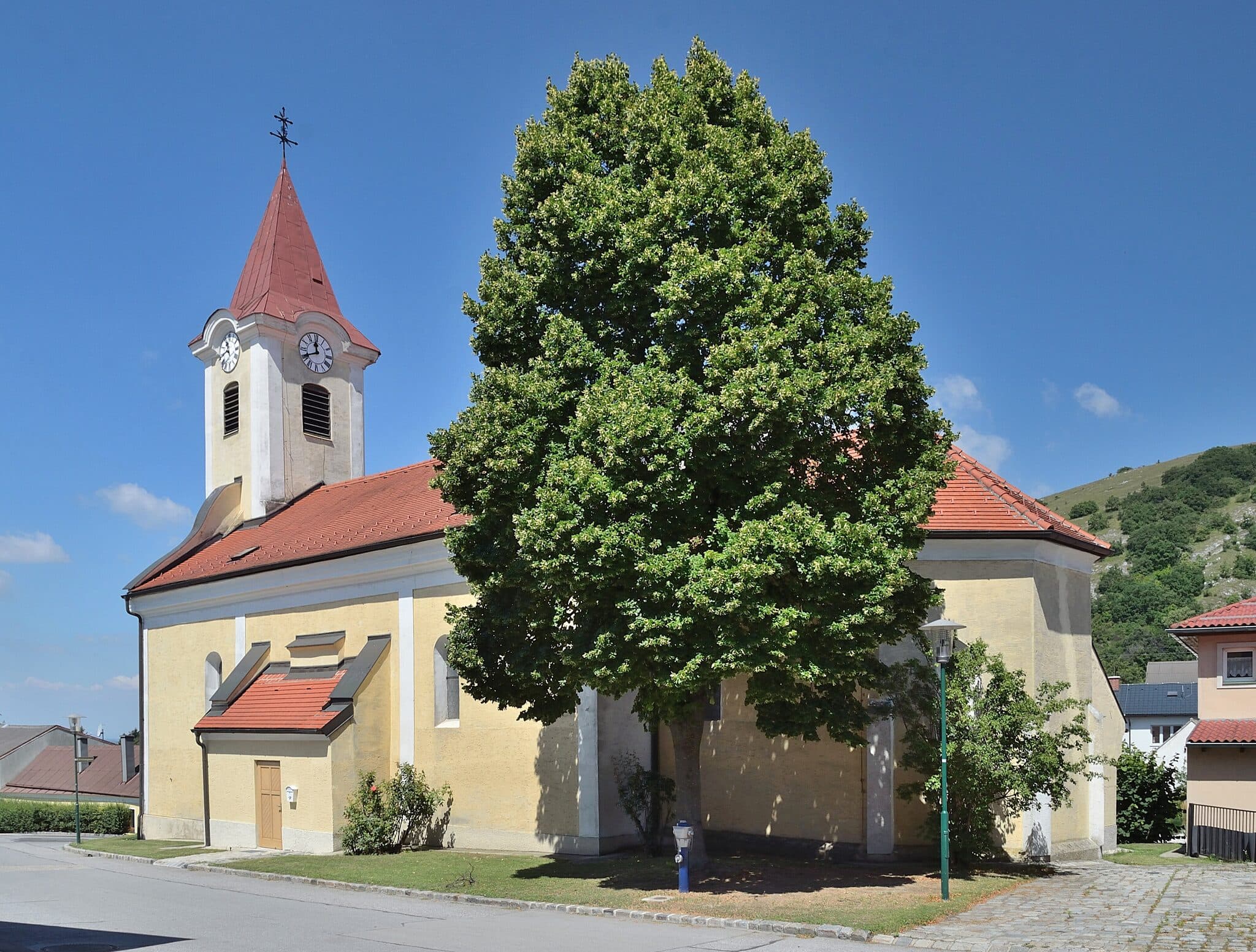 Pfarrkirche Hundsheim mit großem Baum vor blauem Himmel, Ortskontext sichtbar