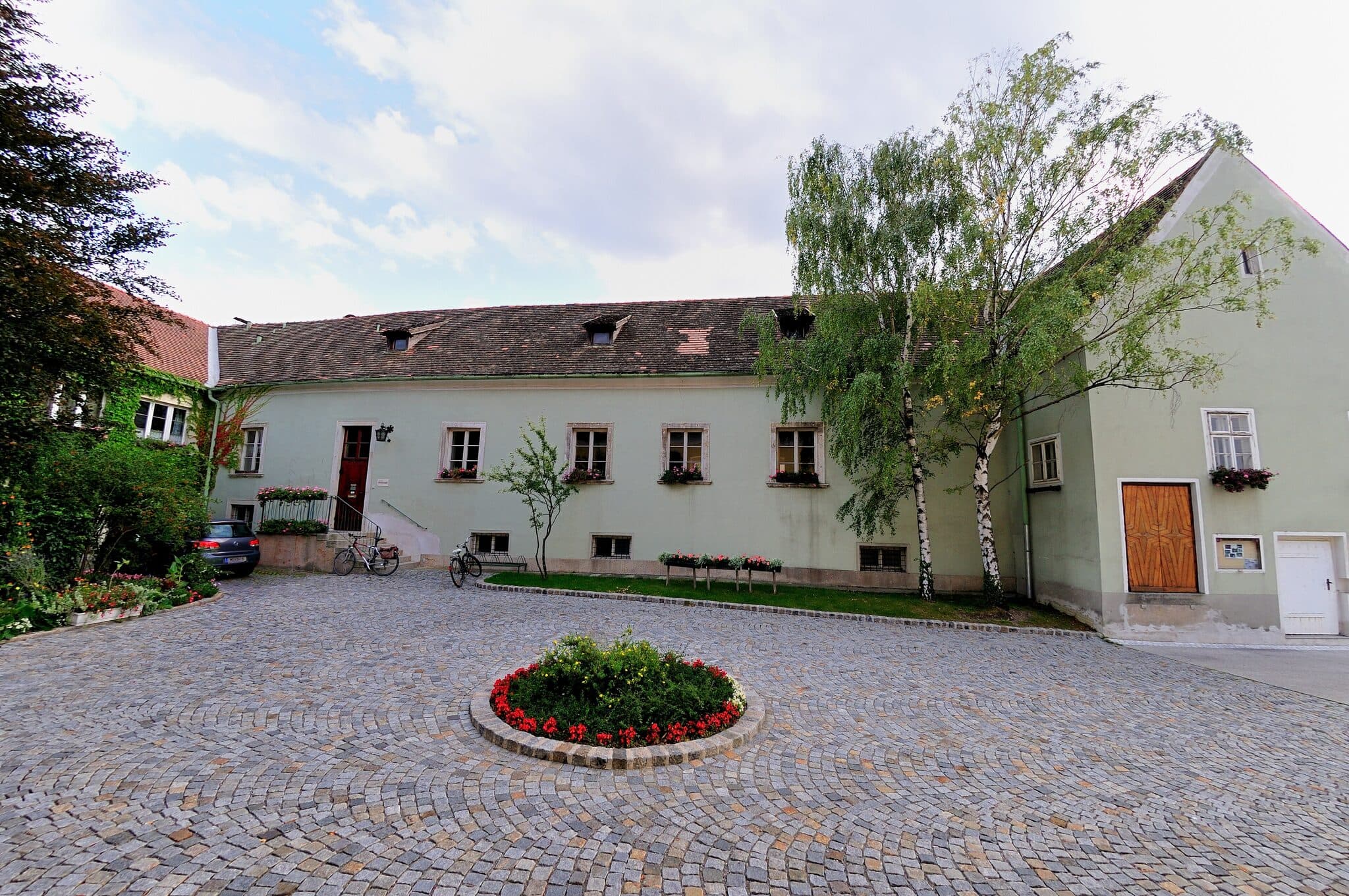 Pfarrhof Leobersdorf - historisches Gebaeude mit Kopfsteinpflaster-Hof, Blumenbeet und Baeumen