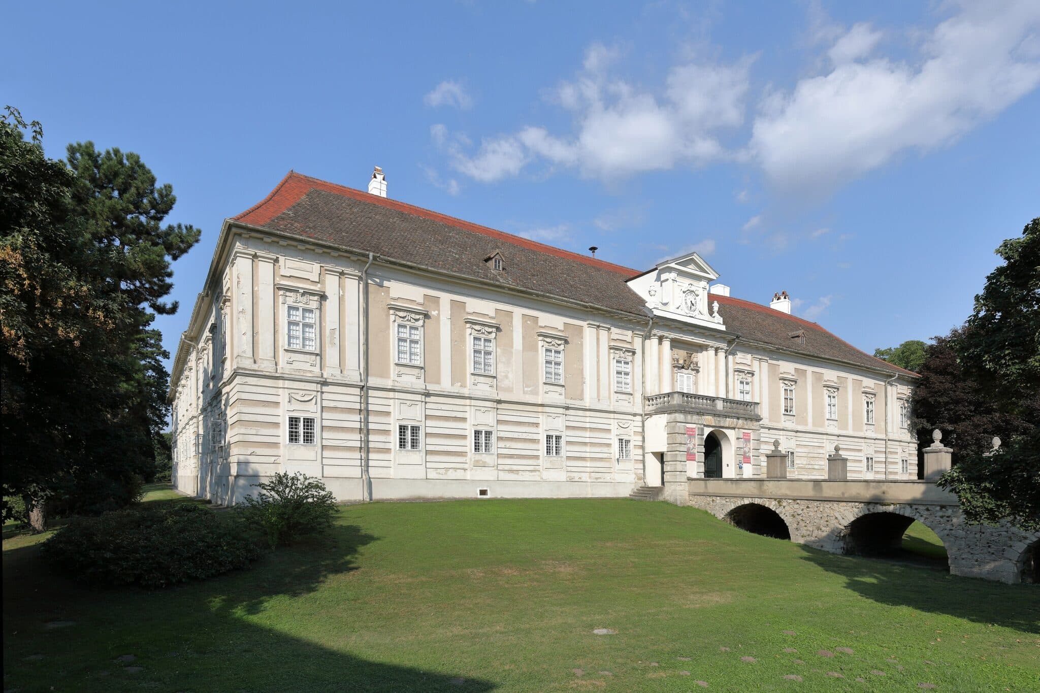 Schloss Harrach in Rohrau - barocke Schlossfassade ueber gepflegtem Rasen mit kleiner Steinbruecke und blauem Himmel