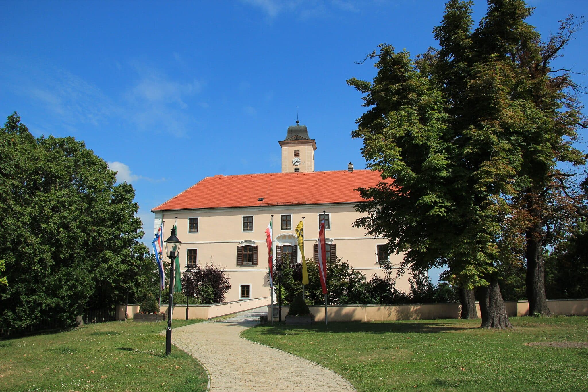 Schloss Vösendorf mit Turm, rotem Dach und Fahnen, blauer Himmel, Rasen, Bäume und einladender Weg – voesendorf